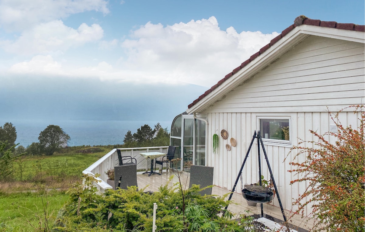 A sunny terrace wraps around the side of the holiday home, featuring a glass conservatory and a swing chair. The view extends toward the fjord, with greenery surrounding the area, creating a serene outdoor space for relaxation.