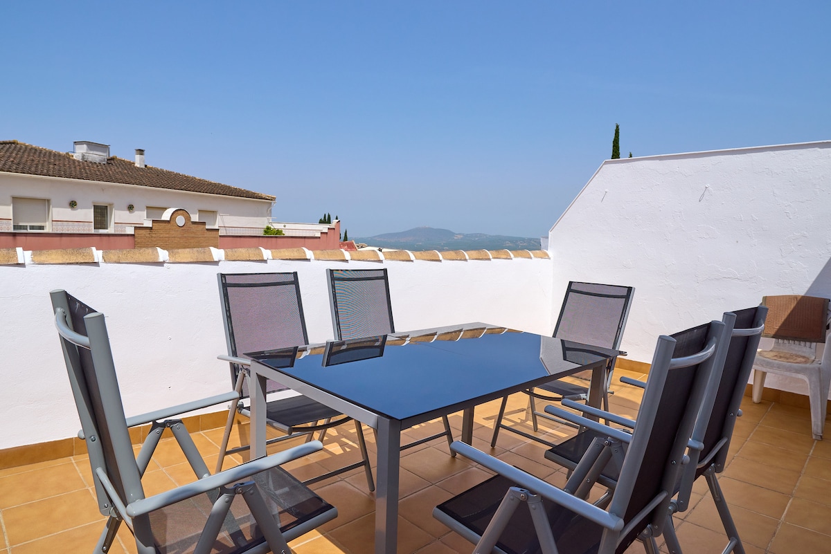 An outdoor terrace features a glass-top table surrounded by six modern chairs. The space offers a clear view of the distant mountains under a blue sky, creating an ideal setting for relaxation or dining al fresco.