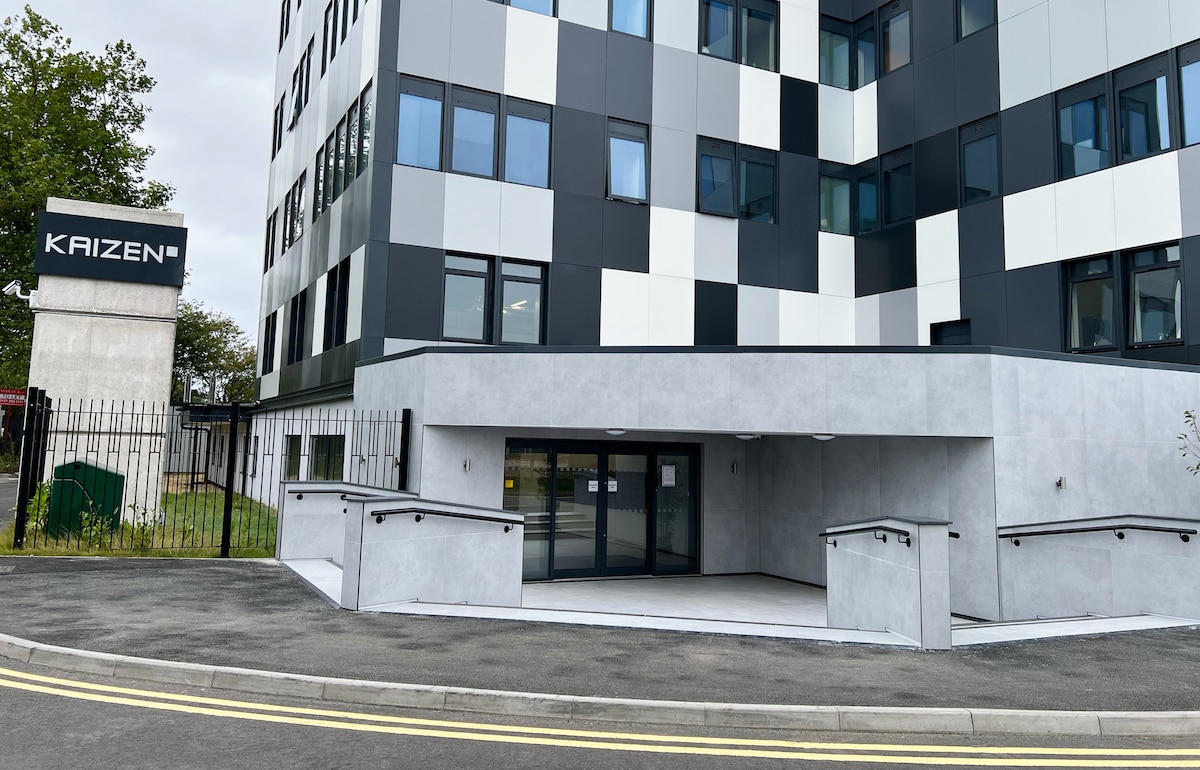 The exterior of the Kaizen building presents a modern architecture with a distinct combination of black and white panels. A wide entrance is accessible via gently sloping ramps, allowing ease of access. The surrounding area features greenery and a well-maintained pathway.