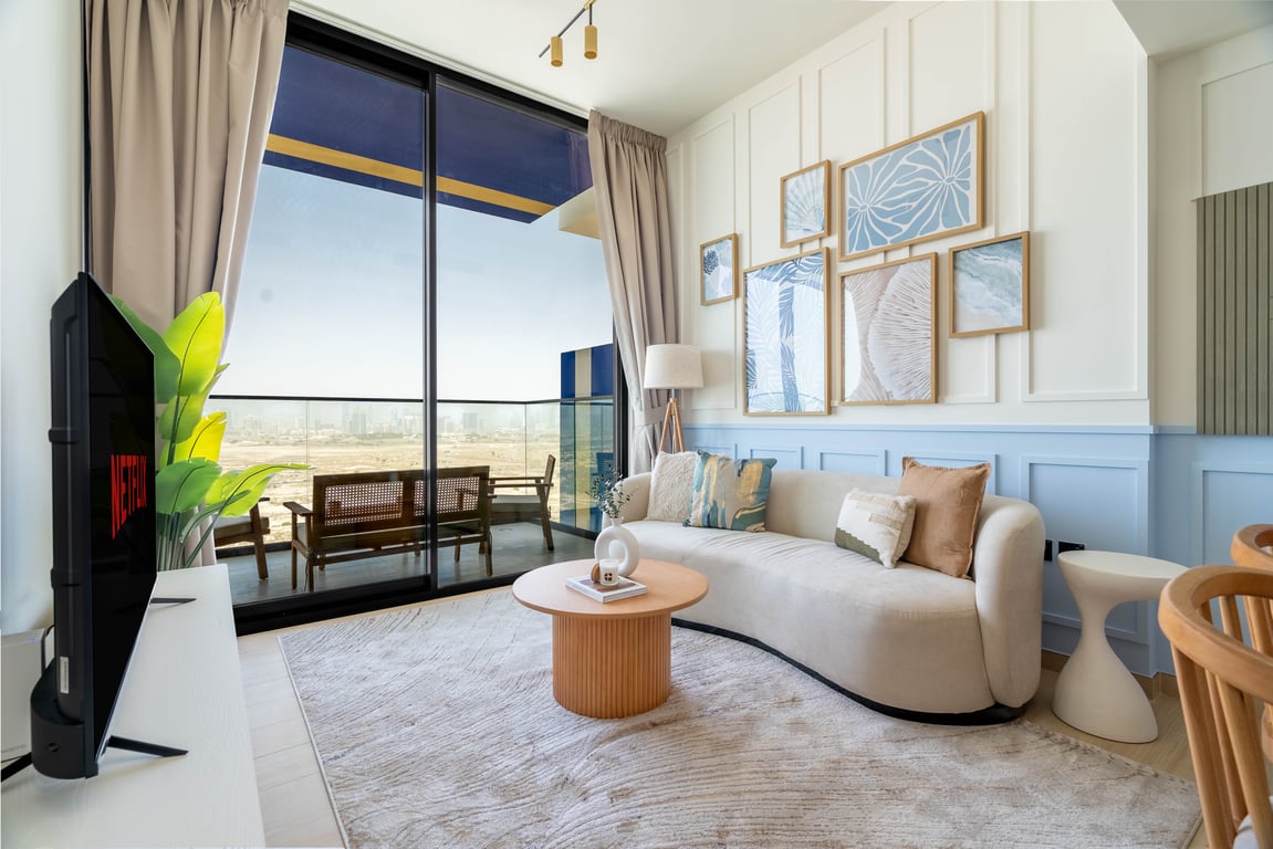 A furnished living area is highlighted by large floor-to-ceiling windows, allowing natural light to fill the space. A curved sofa and circular coffee table create a cozy setting, complemented by decorative wall art and an entertainment unit with a TV, enhancing the modern aesthetic.