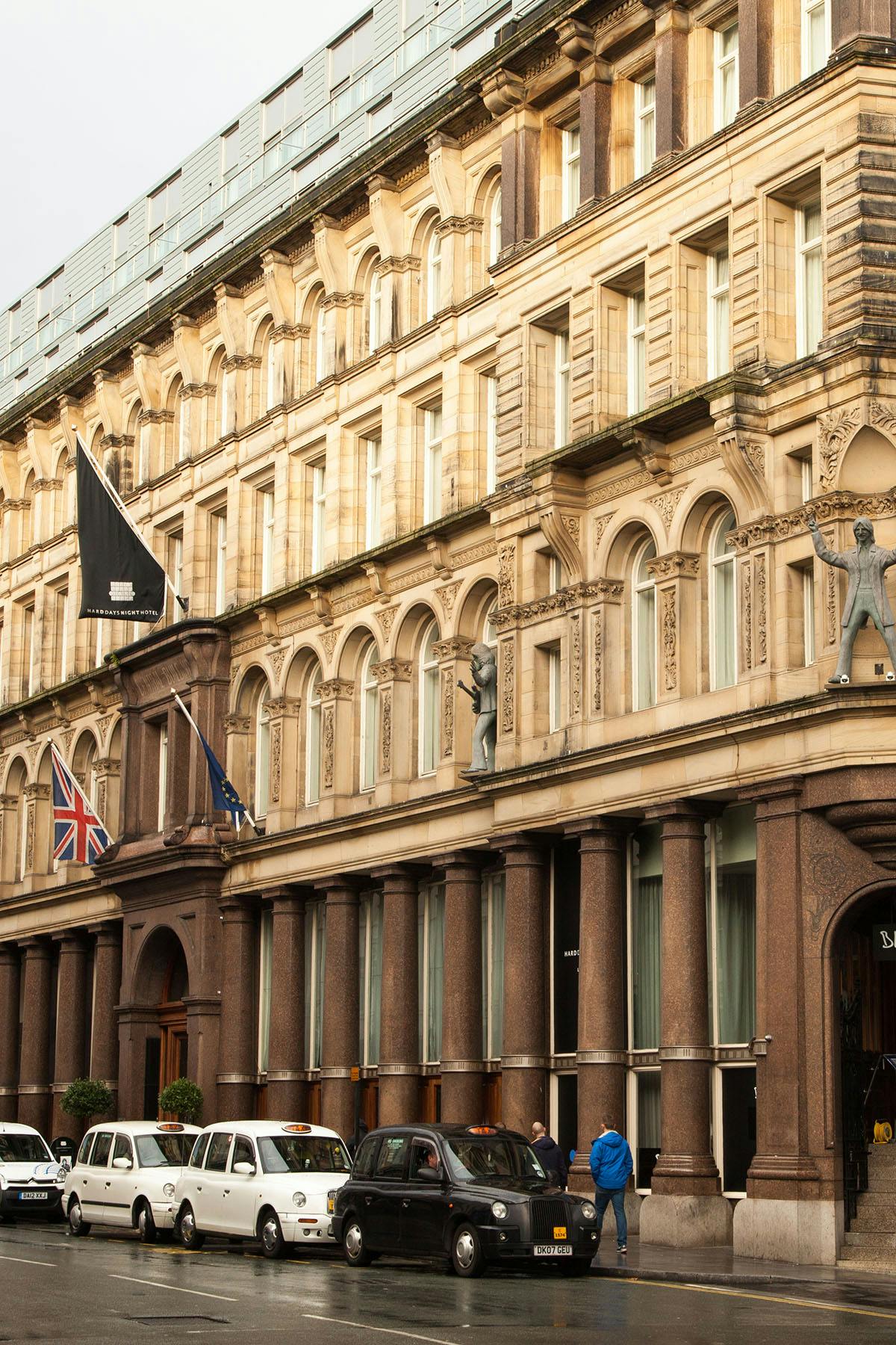 The hotel's elegant exterior is characterized by classic architectural details, including arched windows and ornate stonework. Flags are displayed prominently at the entrance, and a historic building is reflected in the design. The surrounding area features vehicles and pedestrians, adding to the lively atmosphere.