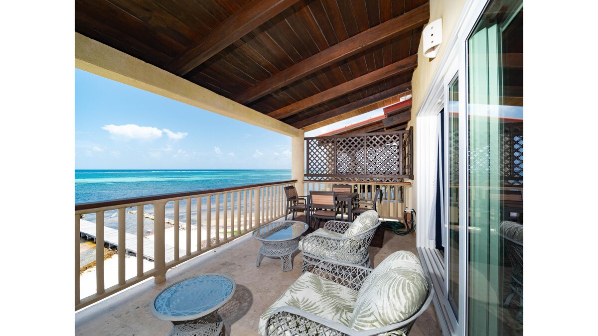 A covered balcony showcases comfortable patio seating arranged around a round table. The ocean stretches beyond, highlighting vibrant turquoise waters and a clear sky. The design features wooden beams overhead, imparting a sense of openness and tranquility.