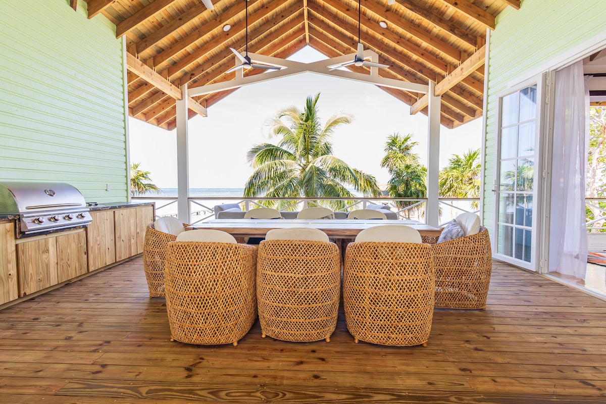 An outdoor seating area features a wooden table surrounded by woven chairs, offering a comfortable spot for gatherings. A barbeque grill is visible to the side, with large doors opening to a view of palm trees and the ocean beyond the spacious deck.