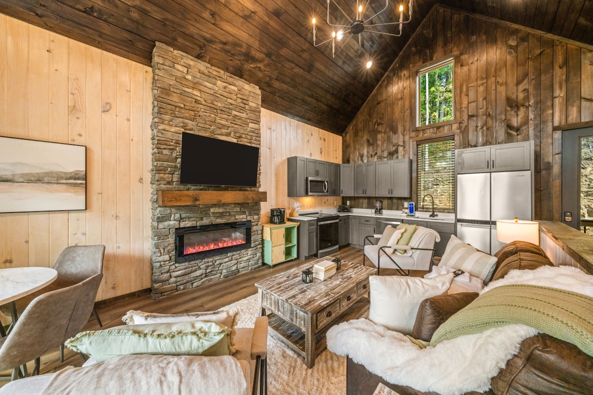 A spacious living area features a stone fireplace serving as a focal point. Modern appliances are arranged in the kitchen, while comfortable seating is positioned around a rustic coffee table. Natural light filters through large windows, highlighting the warm wood interior.