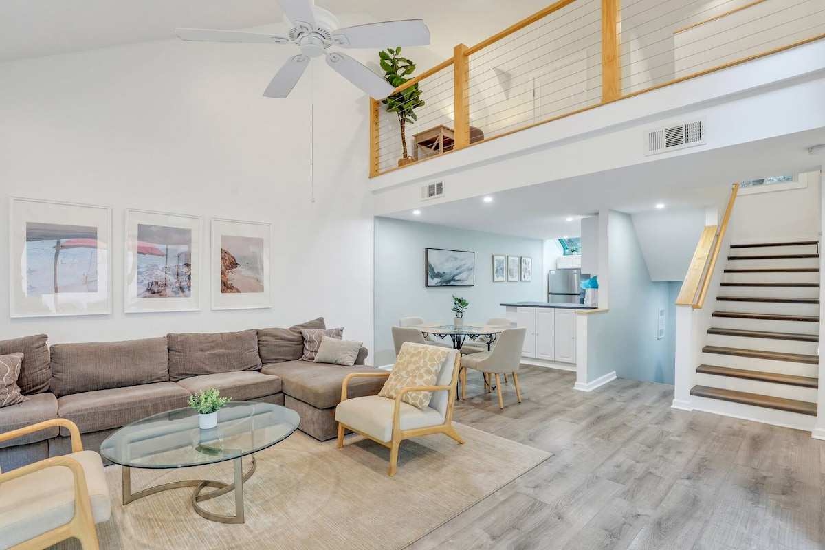 An open living space showcases a comfortable sectional sofa paired with light-colored armchairs. A glass coffee table is centered on a neutral area rug. A dining area is visible in the background, and large windows allow natural light to illuminate the room.
