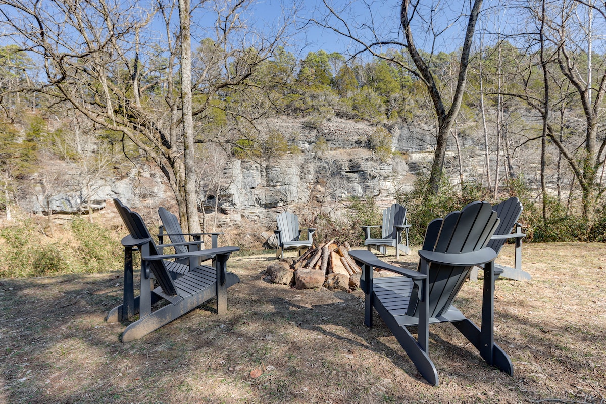 An outdoor seating area is presented with four wooden Adirondack chairs arranged around a central fire pit. A rocky landscape serves as the backdrop, and the space is surrounded by trees, creating a natural ambiance ideal for gatherings.