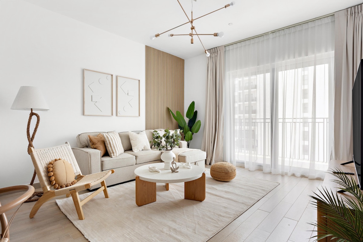 A bright living area is furnished with a cozy sofa, decorative pillows, and a round coffee table. Natural light floods in through the large balcony doors, which are draped with sheer curtains. A decorative lamp and potted plants add inviting touches to the space.