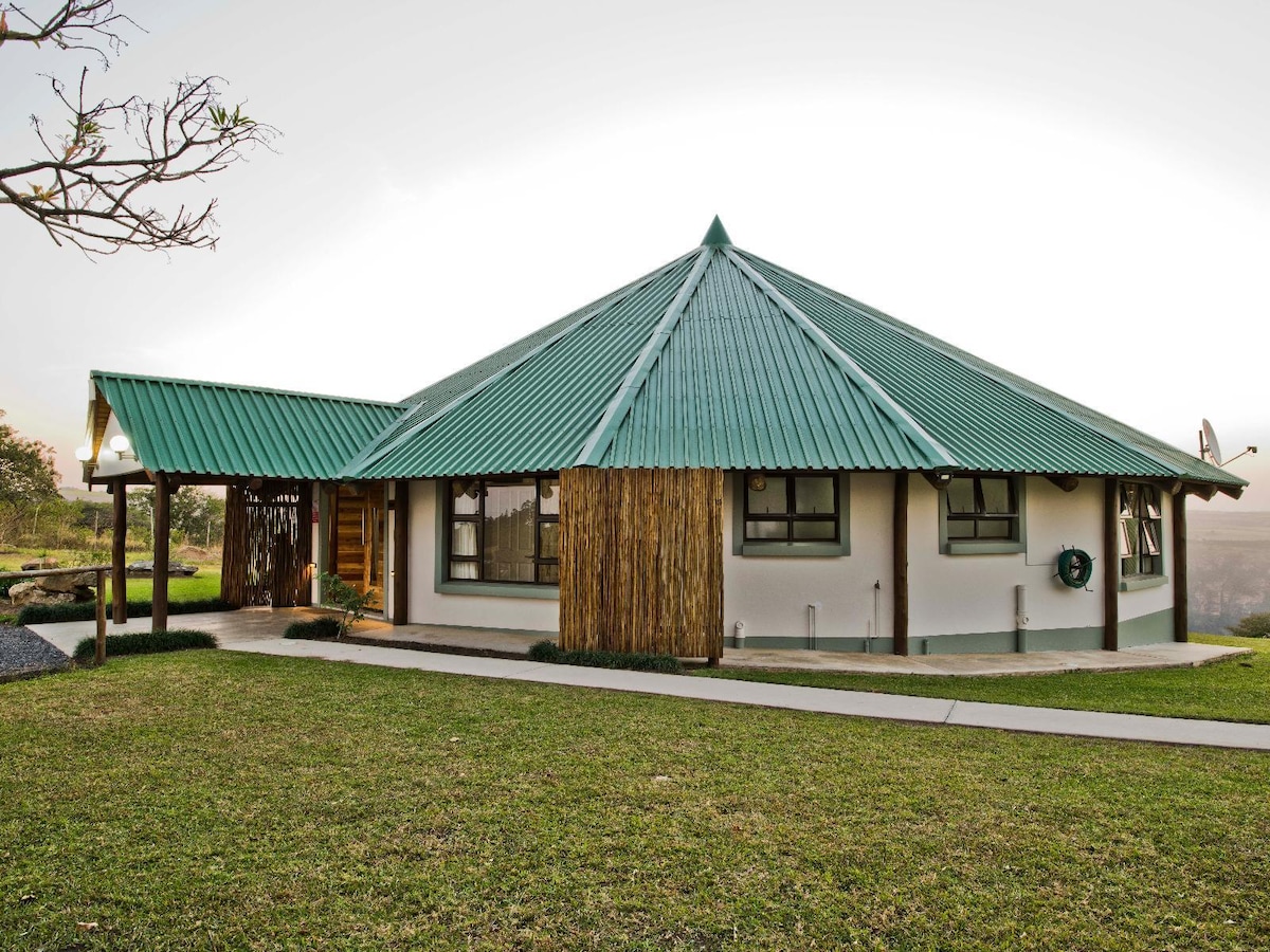 A well-structured house with a distinctive green, triangular roof is set amidst a lush grassy area. Large windows are framed in wood, allowing natural light to illuminate the interior. A concrete pathway leads to the entrance, enhancing accessibility.