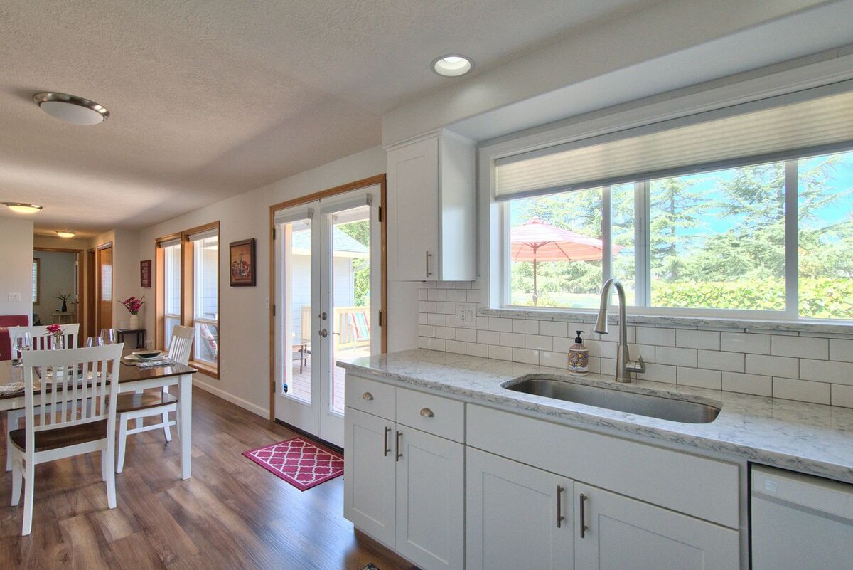 A modern kitchen features sleek white cabinetry and a spacious countertop with a sink. Natural light streams in through large windows, illuminating the open layout that connects to the dining area and outdoor space, where a patio umbrella is visible.