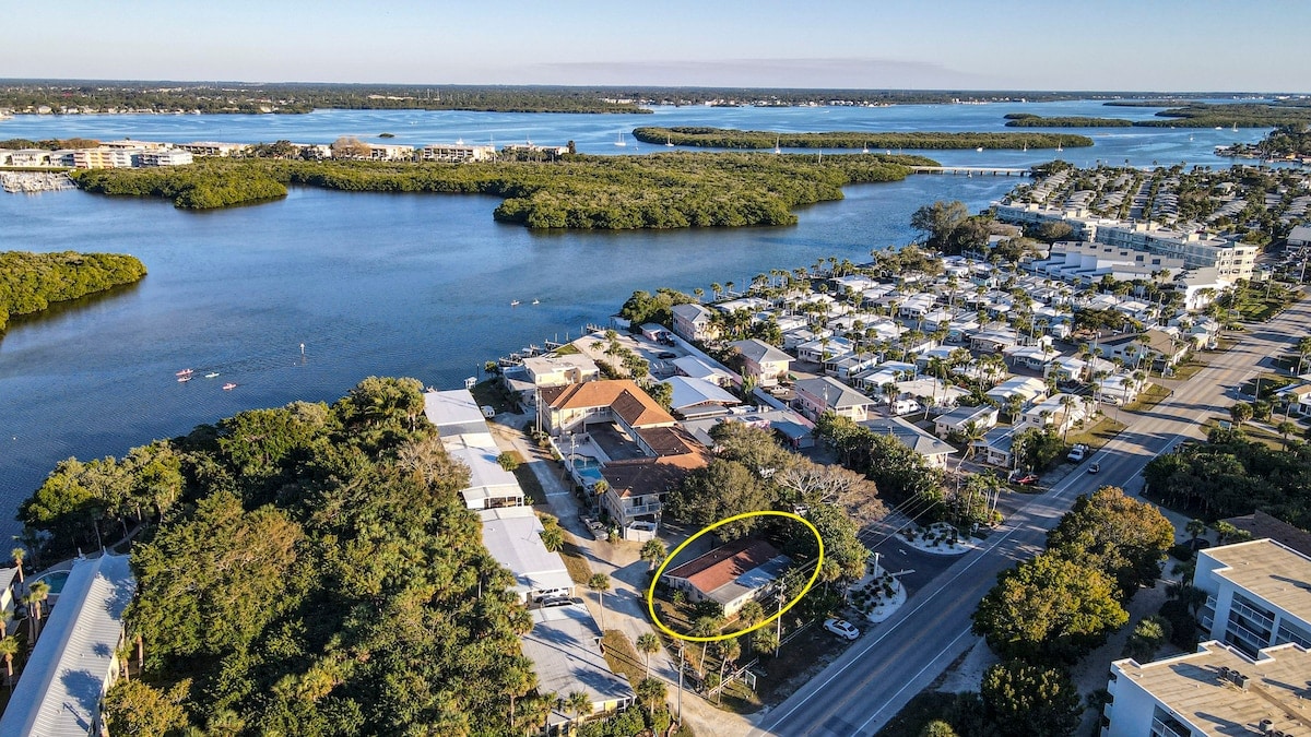 An aerial view captures the surrounding landscape of Manasota Key, showcasing lush green vegetation alongside a winding waterway. The duplex is positioned in the foreground, with nearby residential homes and the road visible, highlighting the coastal proximity and tranquil environment.