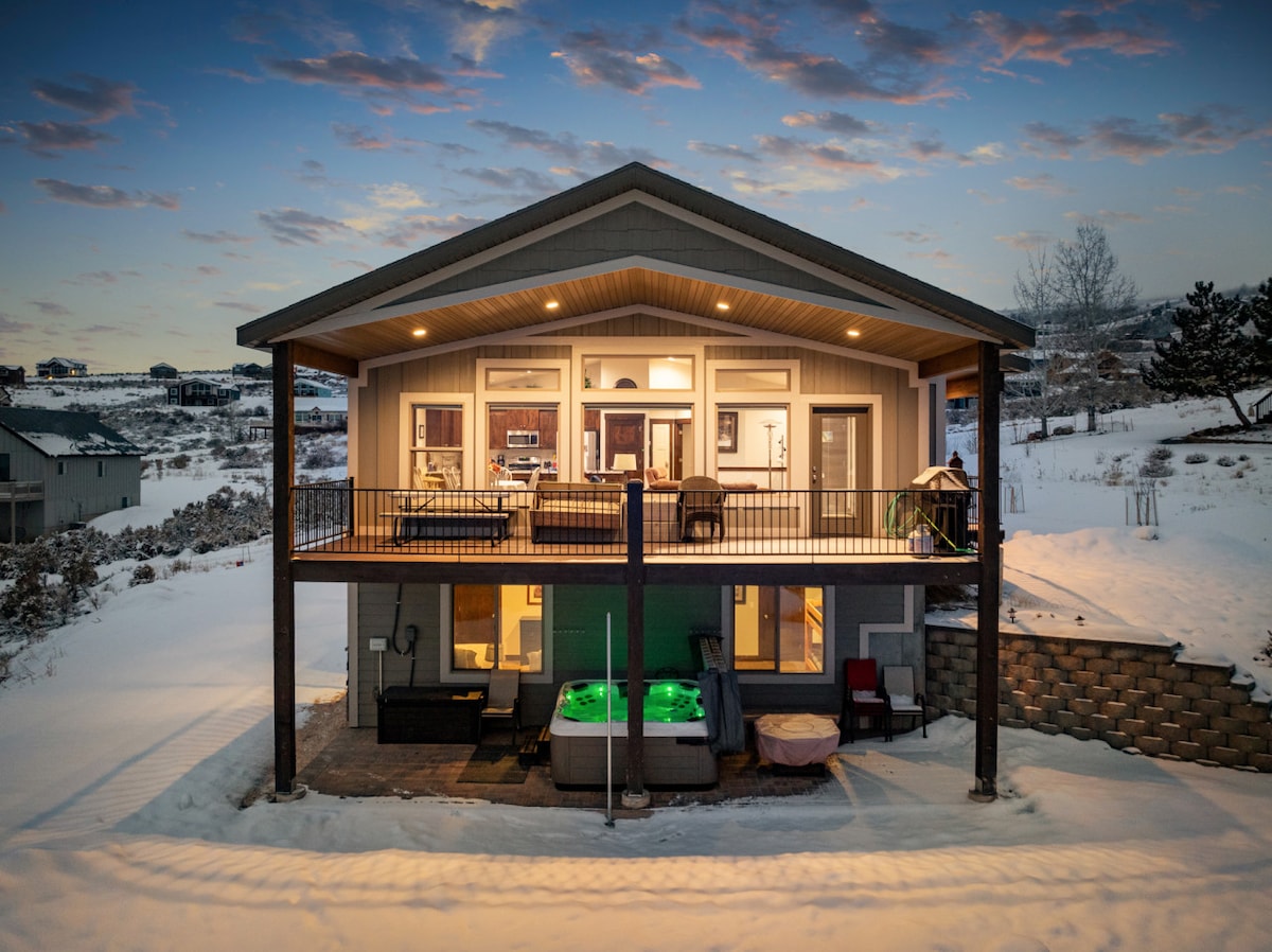 The exterior view of the bungalow features a spacious upper deck with seating and a hot tub, surrounded by snow-covered landscape. Large windows allow visibility into the inviting interior. The structure is framed by a warm sunset sky, enhancing the serene atmosphere.