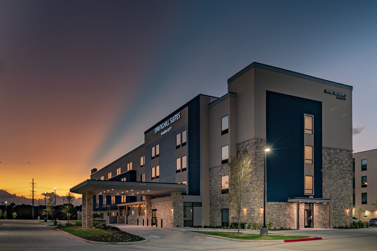 The hotel exterior is captured during dusk, showcasing a contemporary building design. Warm lights illuminate the entrance, while the soft colors of the sunset blend with the darkening sky, creating a calming atmosphere around the landscaped area.