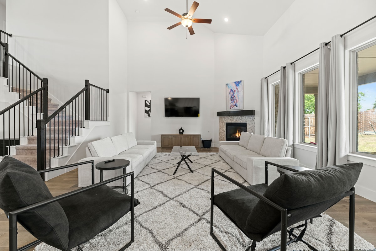 A spacious living room features a high ceiling and large windows allowing natural light. Comfortably arranged seating includes two large sofas and two accent chairs, complemented by a contemporary coffee table. A neutral area rug adds warmth, and a fireplace provides a focal point in the room.