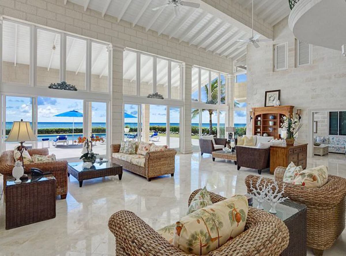 A spacious living area features high ceilings and large glass doors that open to the outdoor space, allowing ample natural light. Comfortable wicker furniture is arranged for relaxation, with decorative pillows adding a touch of detail. The view includes palm trees and the ocean beyond.