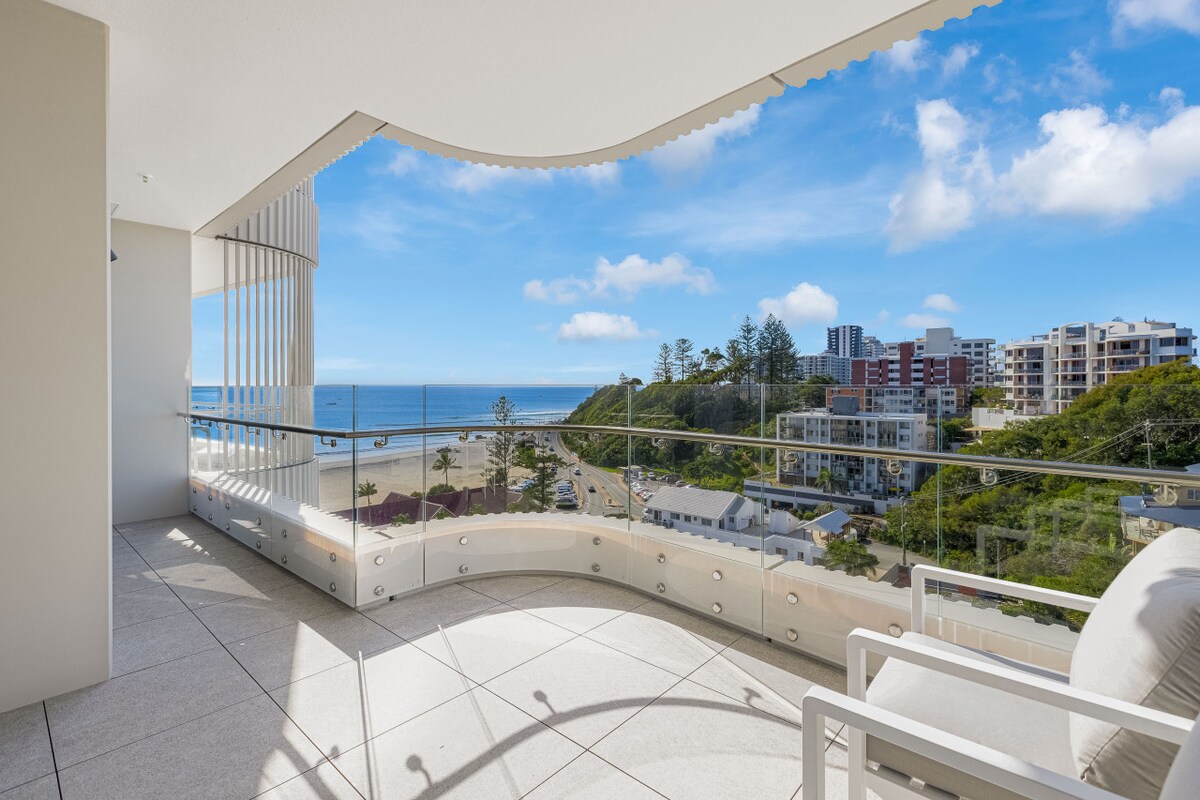 A spacious wrap-around balcony overlooks the ocean and beach, featuring expansive views of Kirra Point. Glass railings enhance visibility, while the sleek tile flooring complements the modern design. A comfortable white chair is positioned to enjoy the serene surroundings.