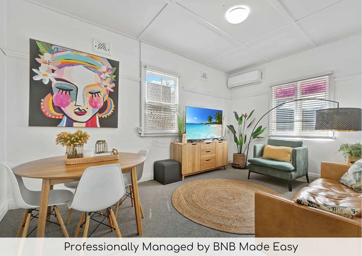 A vibrant living area features a round dining table with white chairs and a wooden cabinet. A large television is positioned next to a cozy green armchair. A colorful mural of a woman’s face adds a touch of artistry, complemented by the soft brown tones of the furnishings.