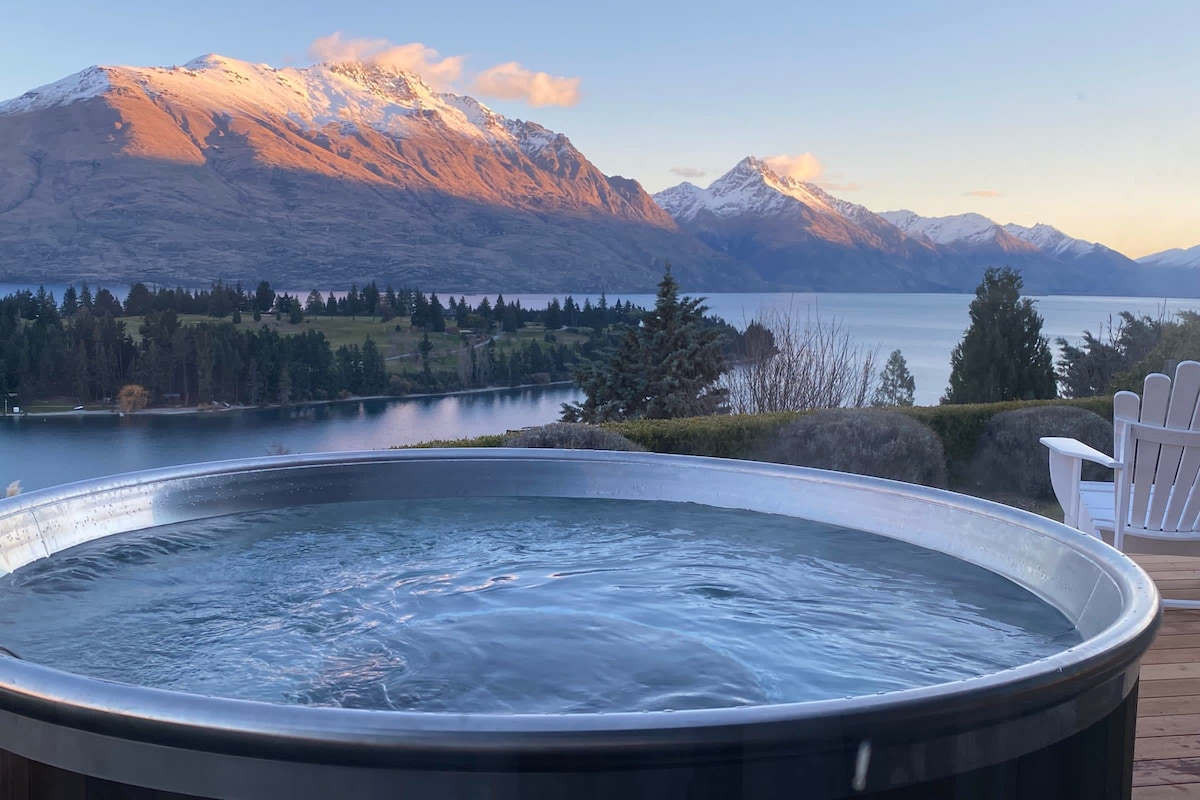 The STOKE Hot Tub is positioned on a wooden deck with a panoramic view of the serene waters of Lake Wakatipu and the surrounding mountains. The snow-capped peaks are illuminated by the soft light of dawn, creating a tranquil and scenic atmosphere.