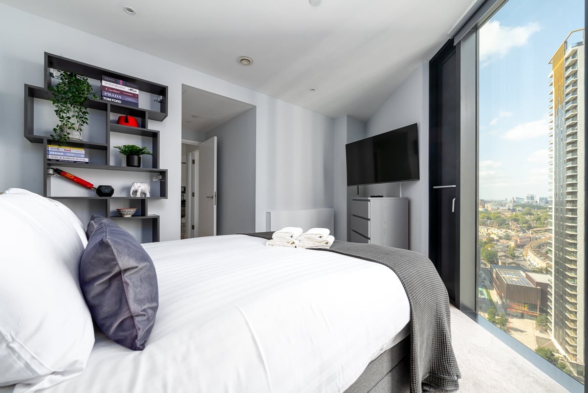 A well-appointed bedroom features a spacious bed adorned with quality linens. A large window offers a view of the city skyline, while a television sits above a minimalistic dresser. A bookshelf on the side displays decorative items, adding an element of style.