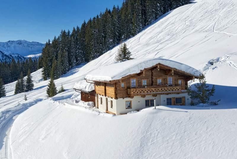 A cozy holiday home is nestled in a snowy landscape, surrounded by evergreen trees under a clear blue sky. The structure features a wooden facade, showcasing traditional architecture. Snow blankets the property and paths, enhancing the serene winter atmosphere.
