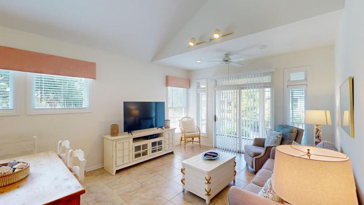 A bright living area features a comfortable seating arrangement with a sofa and armchairs. A large television is centered on a white entertainment unit. Natural light floods in through windows and sliding doors, leading to a balcony. A dining table and chairs are also present.