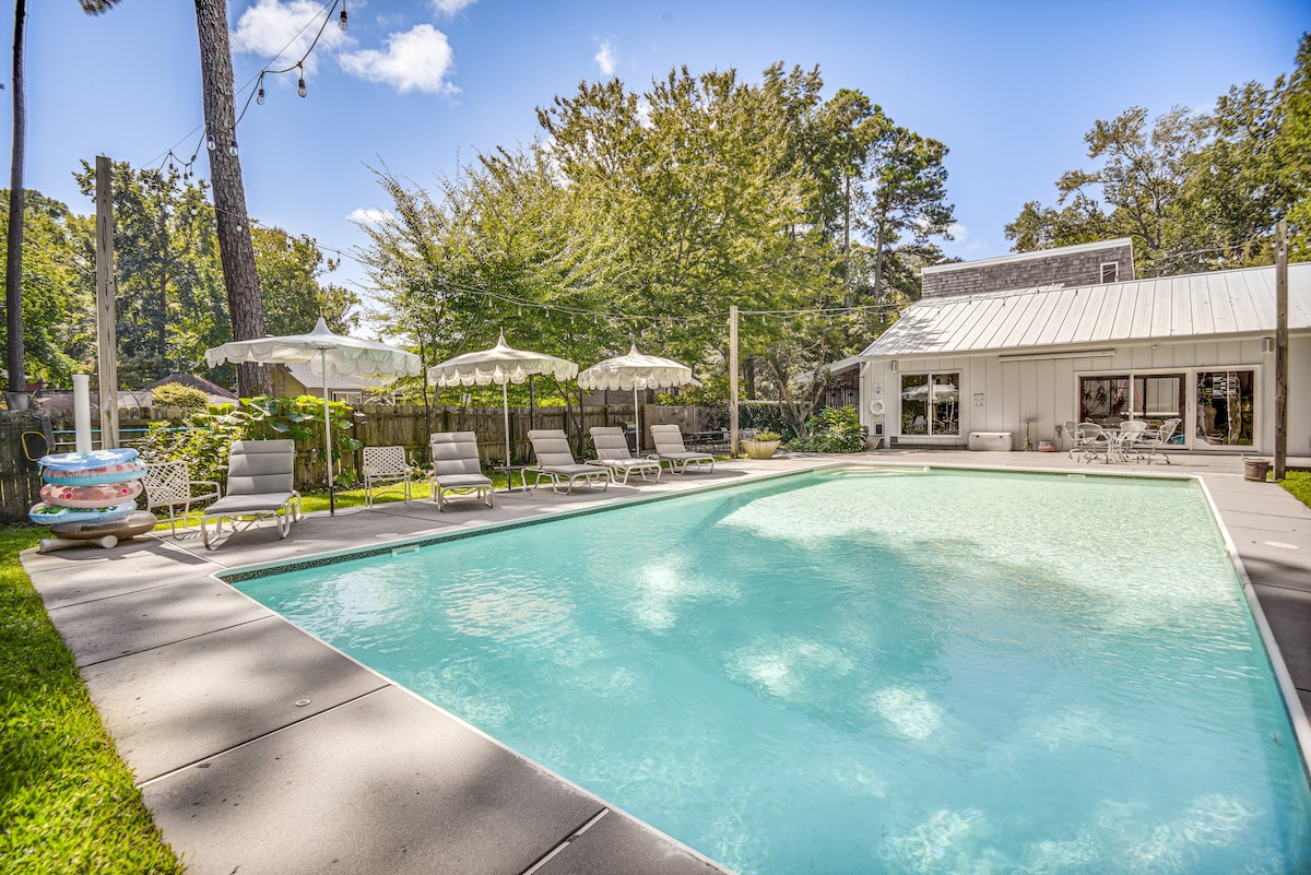 A spacious outdoor pool area is surrounded by sun loungers and shaded umbrellas. Lush greenery is visible in the background, providing a natural ambiance. The pool reflects the blue sky, enhancing the tranquil setting designed for relaxation.