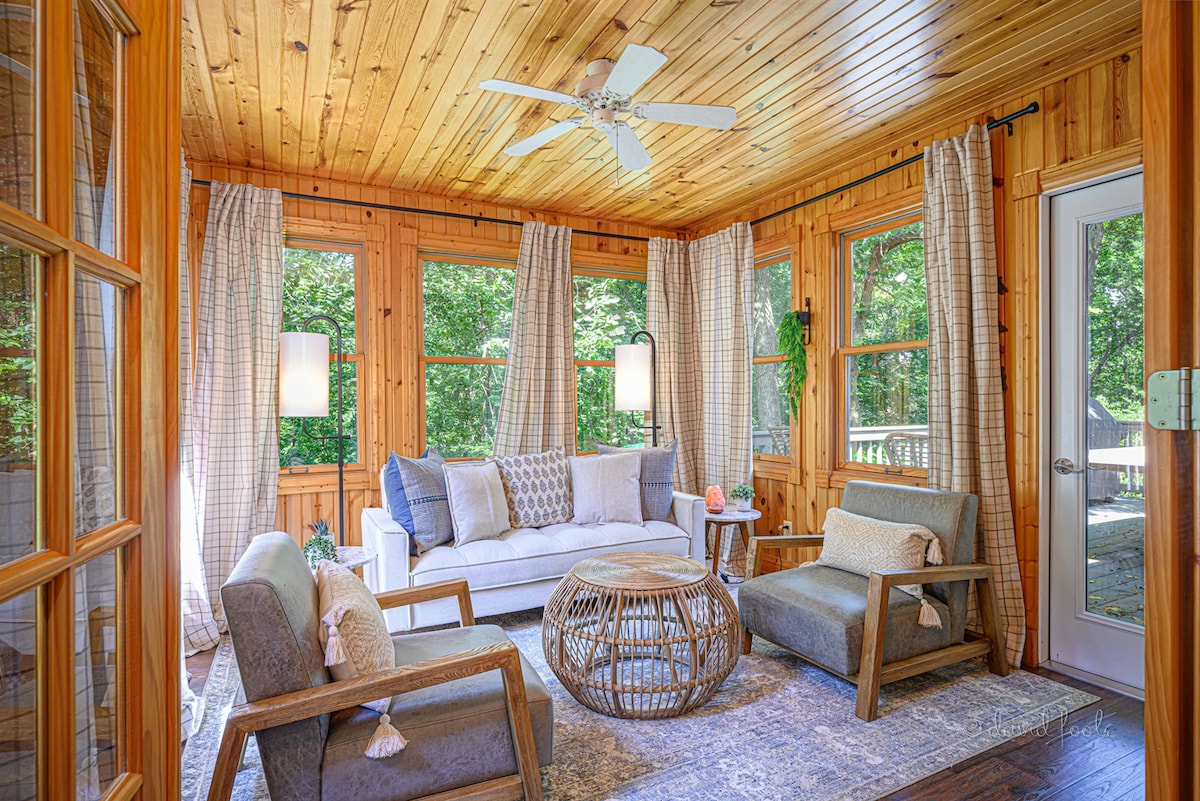A sunlit sunroom features large windows surrounded by wooden walls and ceilings. Two cozy chairs are positioned beside a natural wood coffee table, with a plush sofa offering ample seating. Light-colored curtains softly frame the windows, adding a serene touch to the relaxed atmosphere.