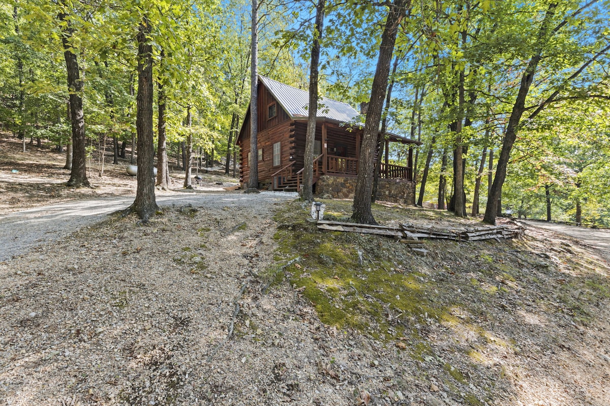A log cabin is nestled among tall trees, surrounded by a gravel pathway. The cabin features a sloped roof and a welcoming porch, blending harmoniously with the natural landscape. Sunlight filters through the leaves, illuminating the serene surroundings with a sense of tranquility.
