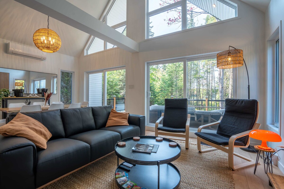 A bright living area showcases large windows that invite natural light, with a view of the surrounding trees. The space features a modern black sofa adorned with plush pillows, paired with two comfortable lounge chairs. A round coffee table holds magazines, and a decorative rug lies beneath.