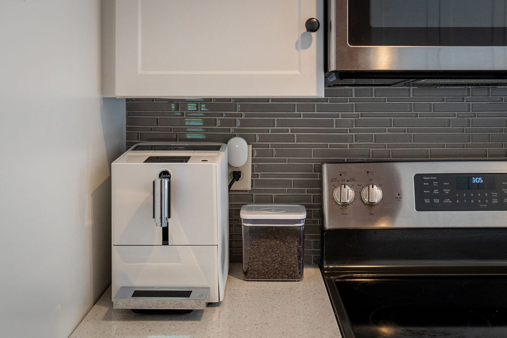 A modern kitchen area features a sleek coffee machine beside a glass container filled with coffee beans. A stainless-steel microwave and stove are also visible, complemented by a contemporary backsplash of gray tiles.