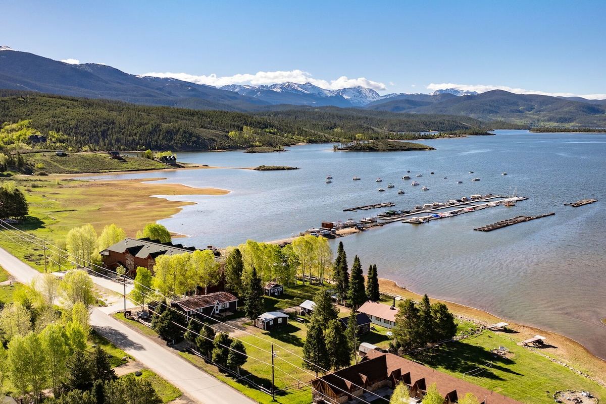 A scenic view of Lake Granby features the expansive blue water surrounded by lush green trees and rugged mountains in the background. The marina is visible with multiple boats docked, and a small stretch of sandy shoreline can be seen along the lake's edge.