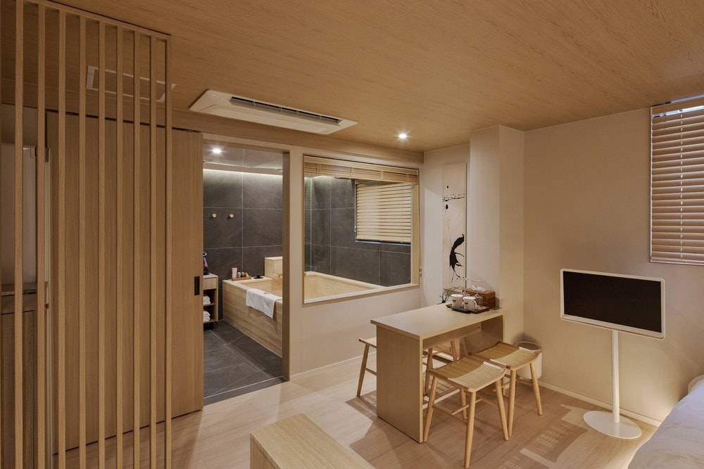 An open room design features light wooden elements, creating a warm ambiance. A cozy table with chairs sits adjacent to a large window. A glass-enclosed bathroom with a bathtub is visible through the partition, enhancing the spacious feel of the area.