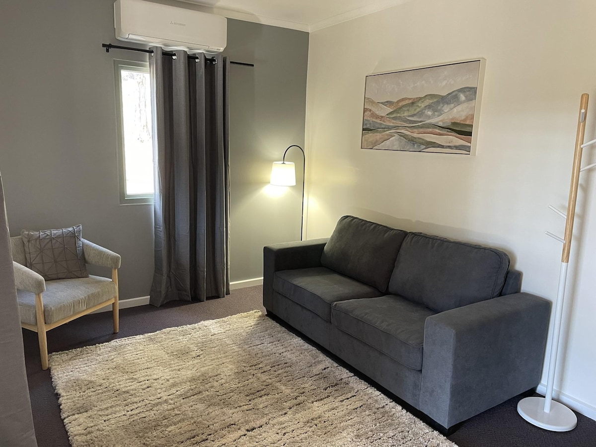 A lounge area features a comfortable sofa, a decorative armchair, and a warm area rug. Natural light enters through a window with soft drapes, while a floor lamp provides additional illumination. A simple landscape artwork decorates the wall, enhancing the room's comfort.
