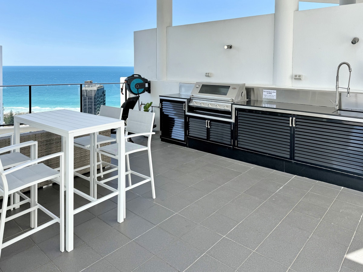The private roof terrace is equipped with a 5-burner gas barbecue and an outdoor kitchen featuring a sink. An 8-seat dining table is positioned beside the ocean view, surrounded by white chairs on a tiled surface, providing ample space for al fresco dining.
