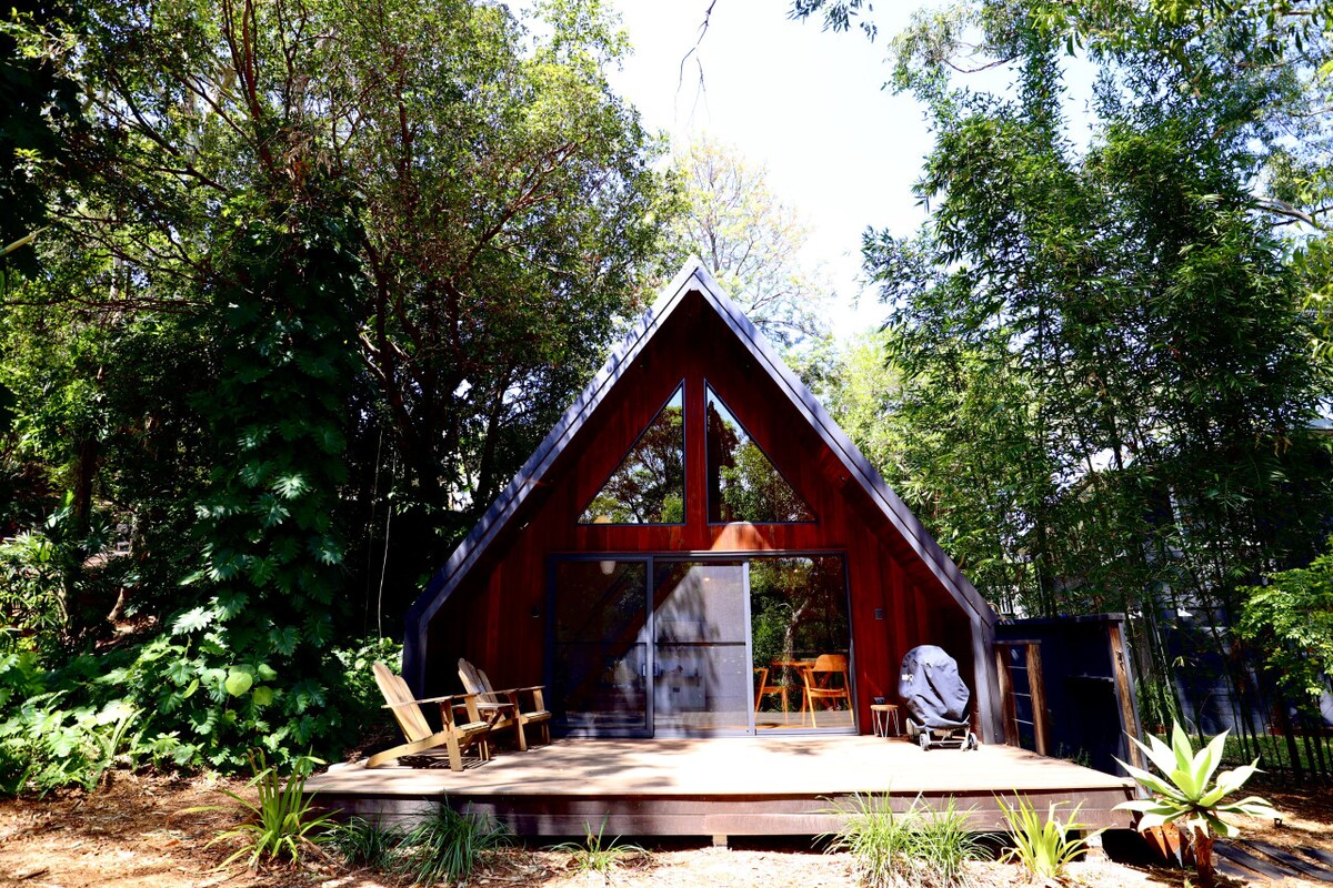 A charming cabin with a distinctive A-frame design is set among lush greenery. The expansive deck features comfortable seating, inviting relaxation while surrounded by nature. Large windows allow natural light to flood the interior, emphasizing the peaceful ambiance of the space.