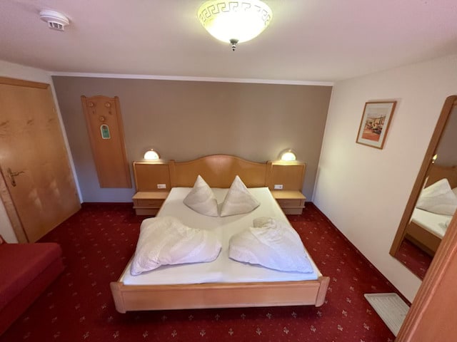 A comfortable sleeping area is featured, showcasing a double bed with two neatly arranged pillows. Soft bedside lighting illuminates the wood accents on the headboard. A full-length mirror and a small seating area with a sofa are visible, enhancing the room's functionality.