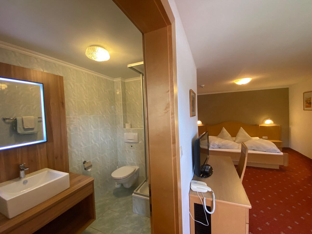A spacious room is visible, featuring a double bed with soft bedding. A bathroom is seen with a modern sink and shower area. A television and wooden furniture complement the room, while warm lighting contributes to a cozy ambiance.