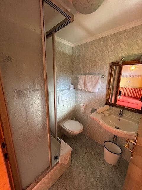 A compact bathroom showcases a glass shower enclosure, a toilet, and a sink with a circular mirror above it. Soft white towels are neatly hung on a rack. Light-colored tile lines the floor and walls, creating a clean and bright environment.