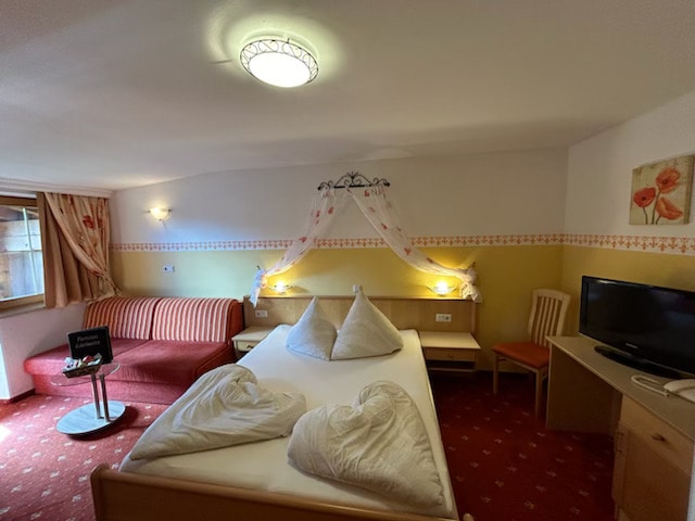 A well-appointed double room features a spacious bed adorned with heart-shaped pillows. A comfy sofa is located next to a window that allows natural light to fill the space. A wooden chair and a television are positioned near the desk, complementing the inviting decor.