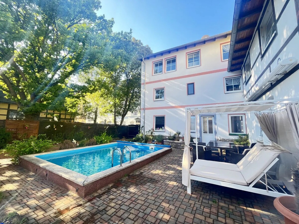 A shared outdoor area features a rectangular pool surrounded by a paved patio. A white lounge bed sits nearby, offering a relaxing spot under the clear blue sky. Lush greenery and trees provide shade and enhance the tranquil environment.
