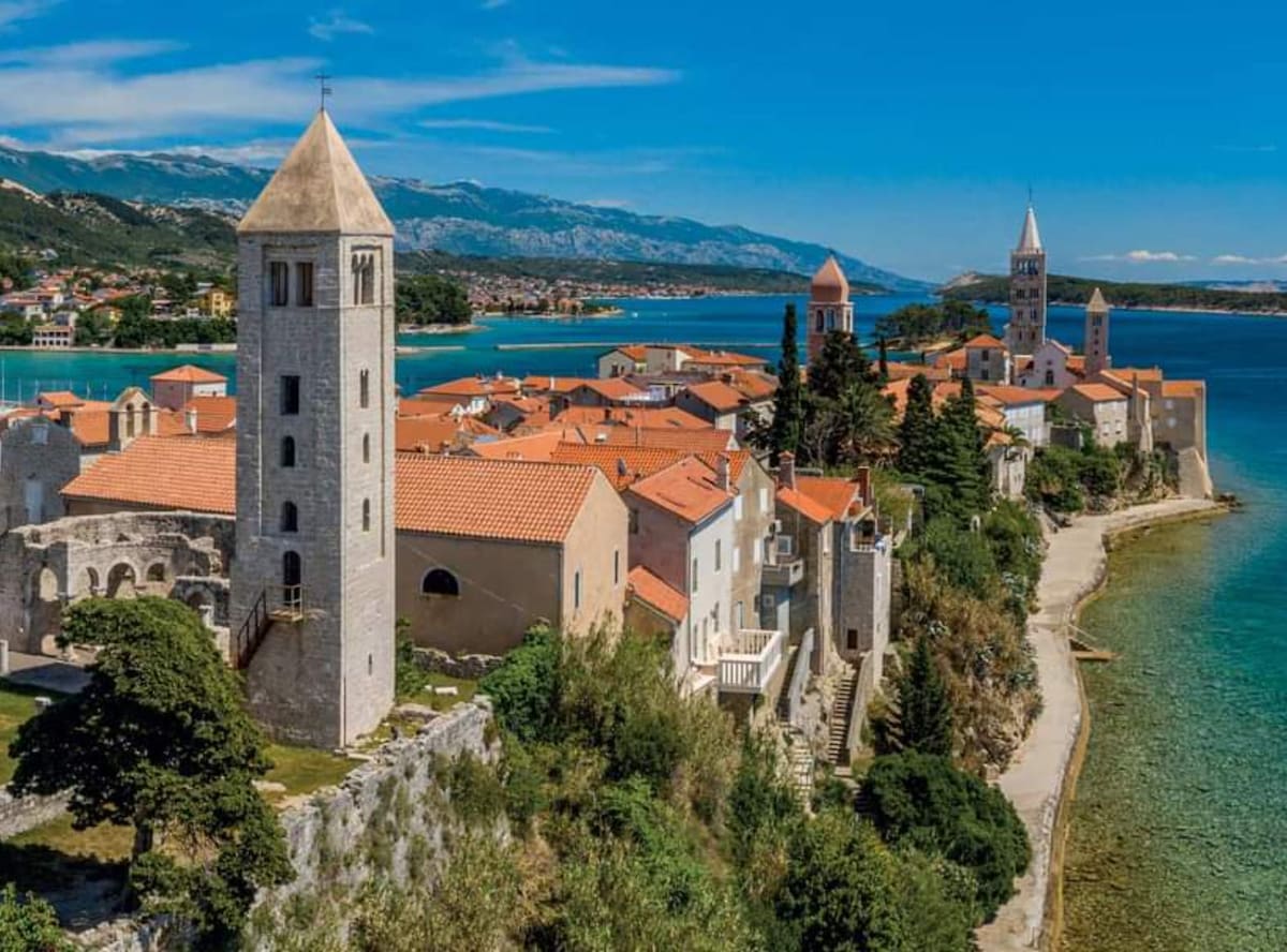 The coastal village is characterized by historic stone towers and buildings with orange-tiled roofs, set against a backdrop of lush green hills and a clear blue sky. Calm waters reflect the scenery, while a winding shoreline invites exploration.