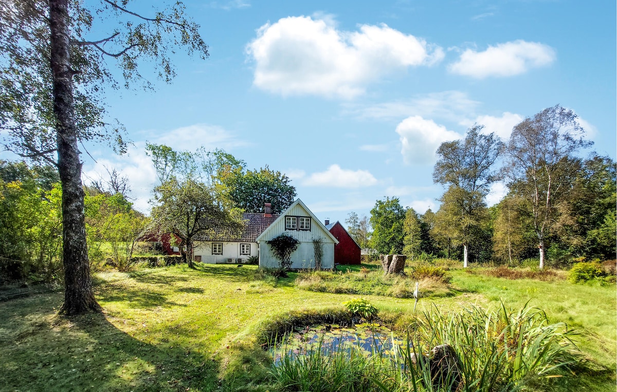 A charming holiday home is nestled within lush greenery, surrounded by trees and a well-kept lawn. A tranquil pond is visible in the foreground, complemented by natural plant life. The house features a quaint red structure alongside the main building, creating a picturesque view.