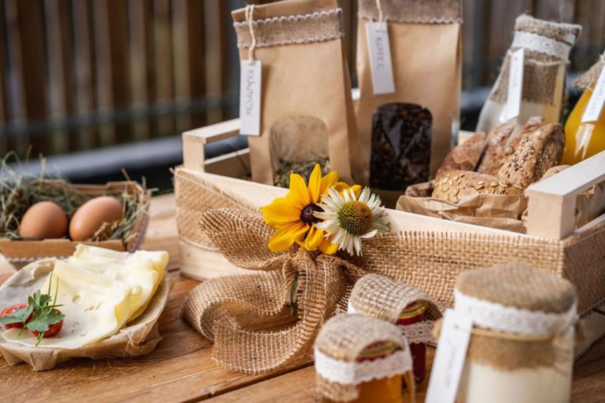 A rustic wooden basket filled with various local products is displayed on a wooden surface. Items include jars of preserves, bags of tea or herbs, baked goods, fresh eggs, and slices of cheese, all adorned with a sunflower and burlap ribbon for a charming presentation.