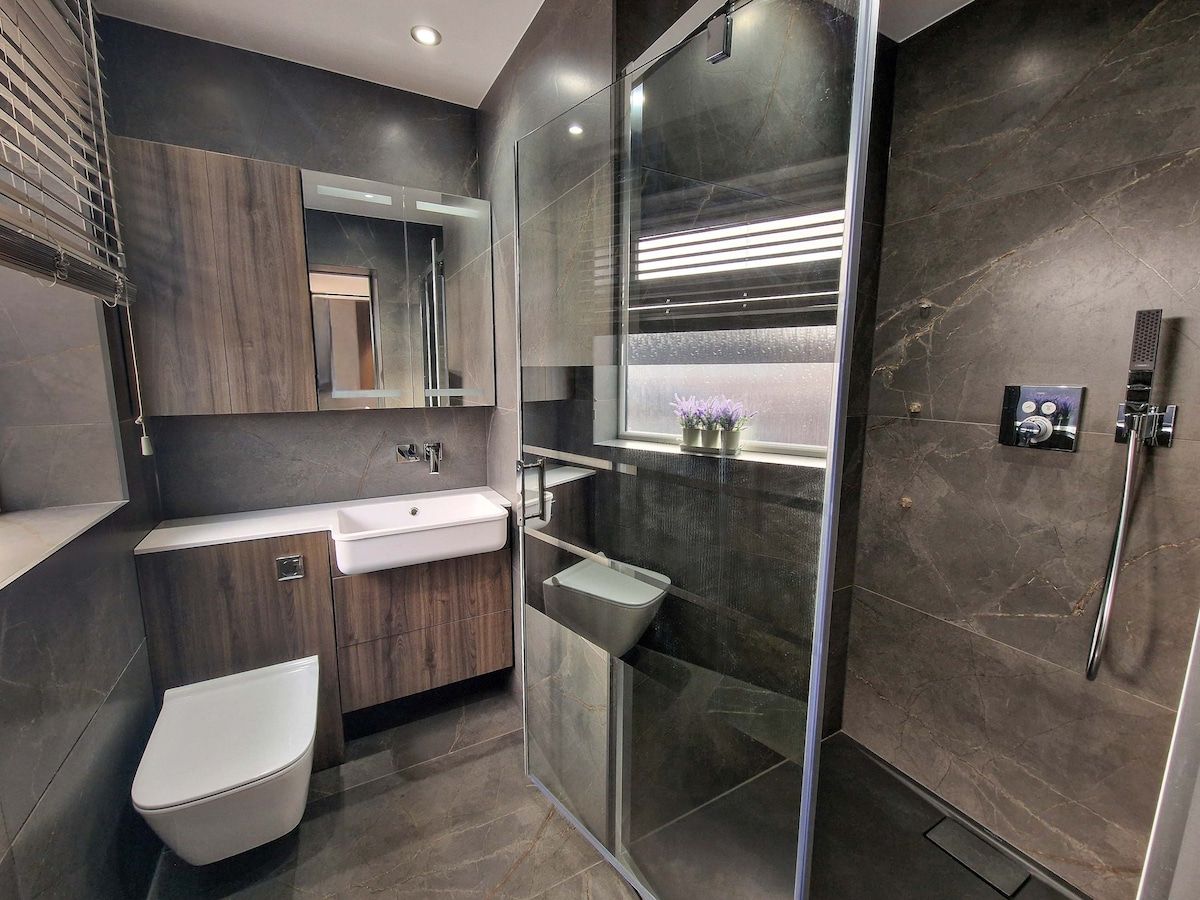 The modern bathroom features a glass-enclosed shower and a sleek, wall-mounted toilet. Dark tile flooring complements the wood accents on the vanity and cabinetry. A large mirror reflects the natural light and flowers placed on the countertop, enhancing the space's contemporary design.