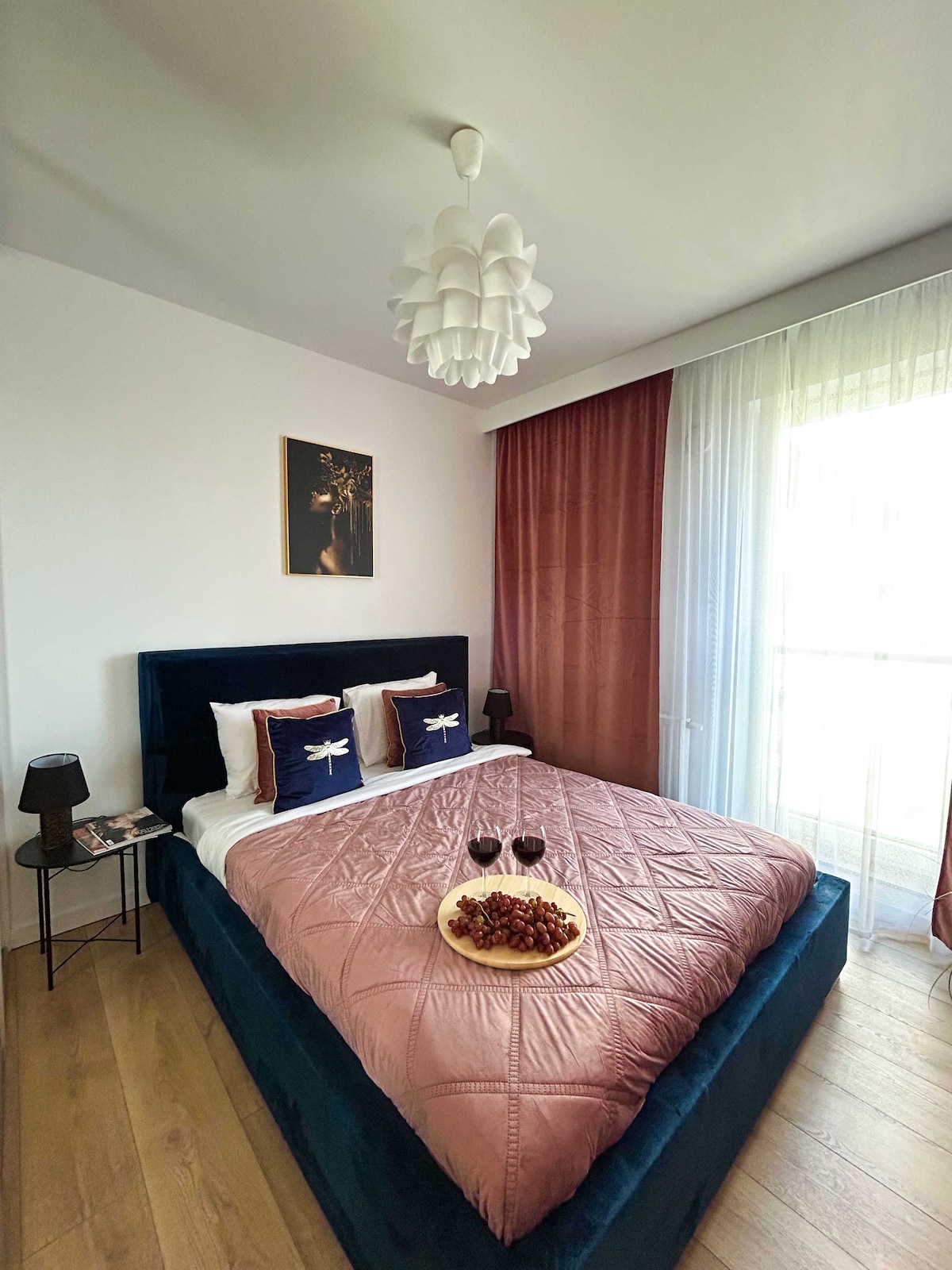 A cozy bedroom features a spacious bed with a soft pink coverlet and decorative pillows. Natural light filters through a sheer curtain, illuminating the space. A small side table holds a decorative plate with fruits and two glasses, creating a welcoming ambiance.