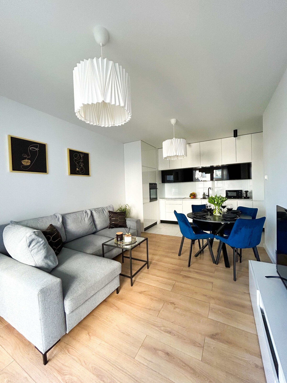 A modern living area features a light grey sofa accompanied by a dark coffee table. A dining table with blue chairs is set against a stylish black kitchen, which is equipped with modern appliances. Two decorative light fixtures hang from the ceiling, complementing the space.