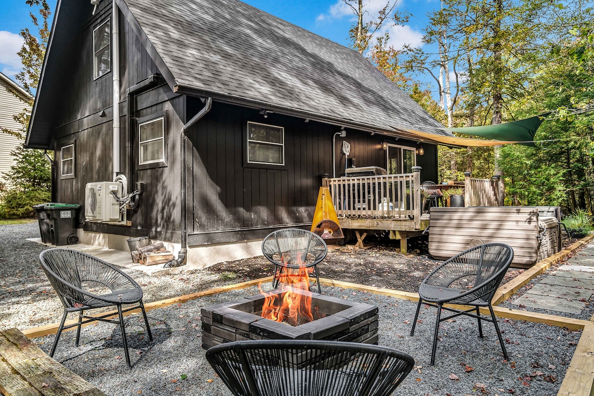 An outdoor gathering space features a fire pit surrounded by four chairs, positioned on gravel. The sleek, dark exterior of the house is complemented by a wooden deck and a green awning, with trees providing a natural backdrop.