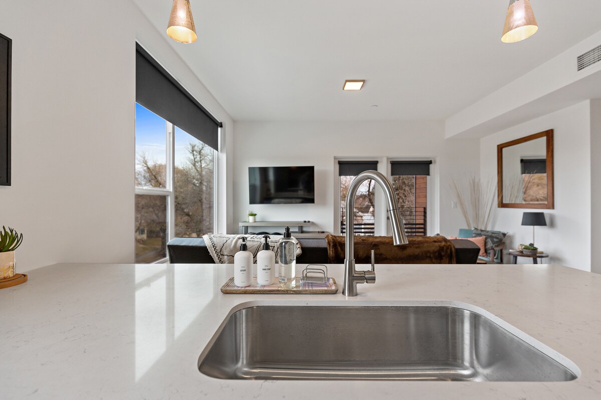 A modern kitchen space is presented, featuring a sleek sink and countertop. In the background, a living area with a soft couch and wall-mounted TV can be seen. Large windows invite natural light, while pendant lighting adds a warm touch.