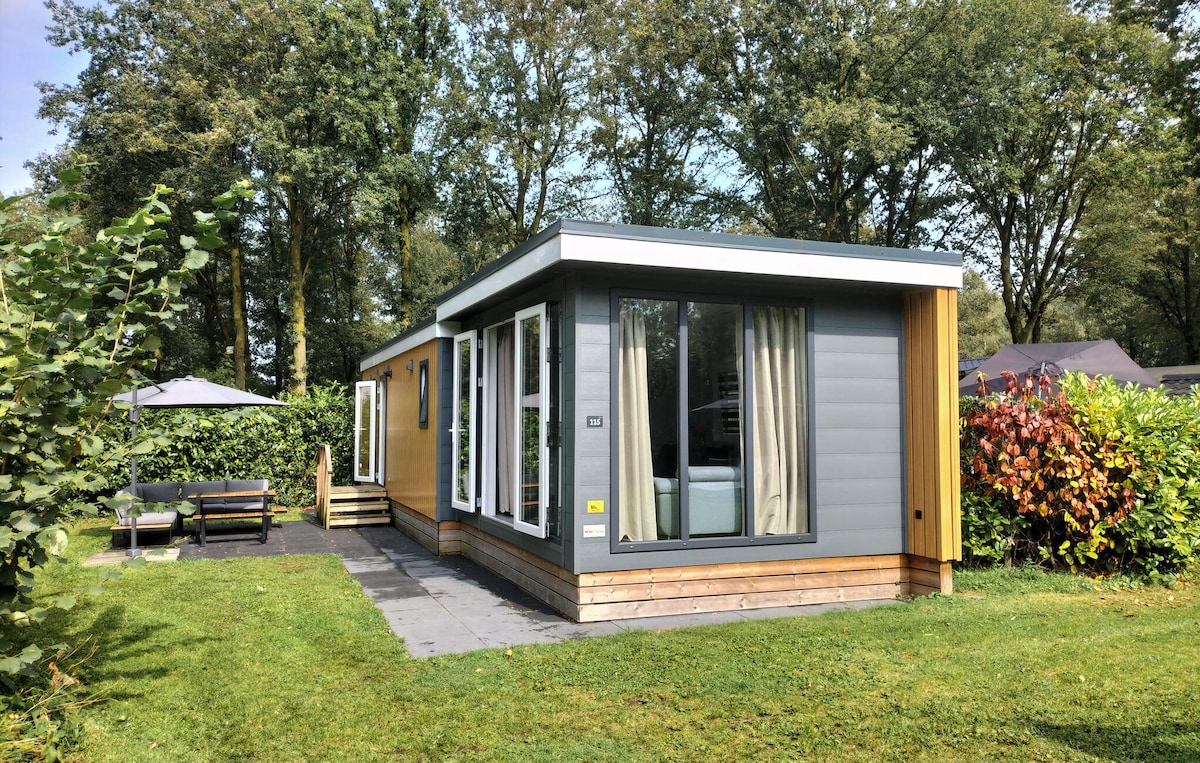The chalet is presented in a modern design with a combination of grey and wooden siding. Large windows allow ample natural light to enter, framed by light curtains. A patio area features seating and is surrounded by lush greenery and shrubs.