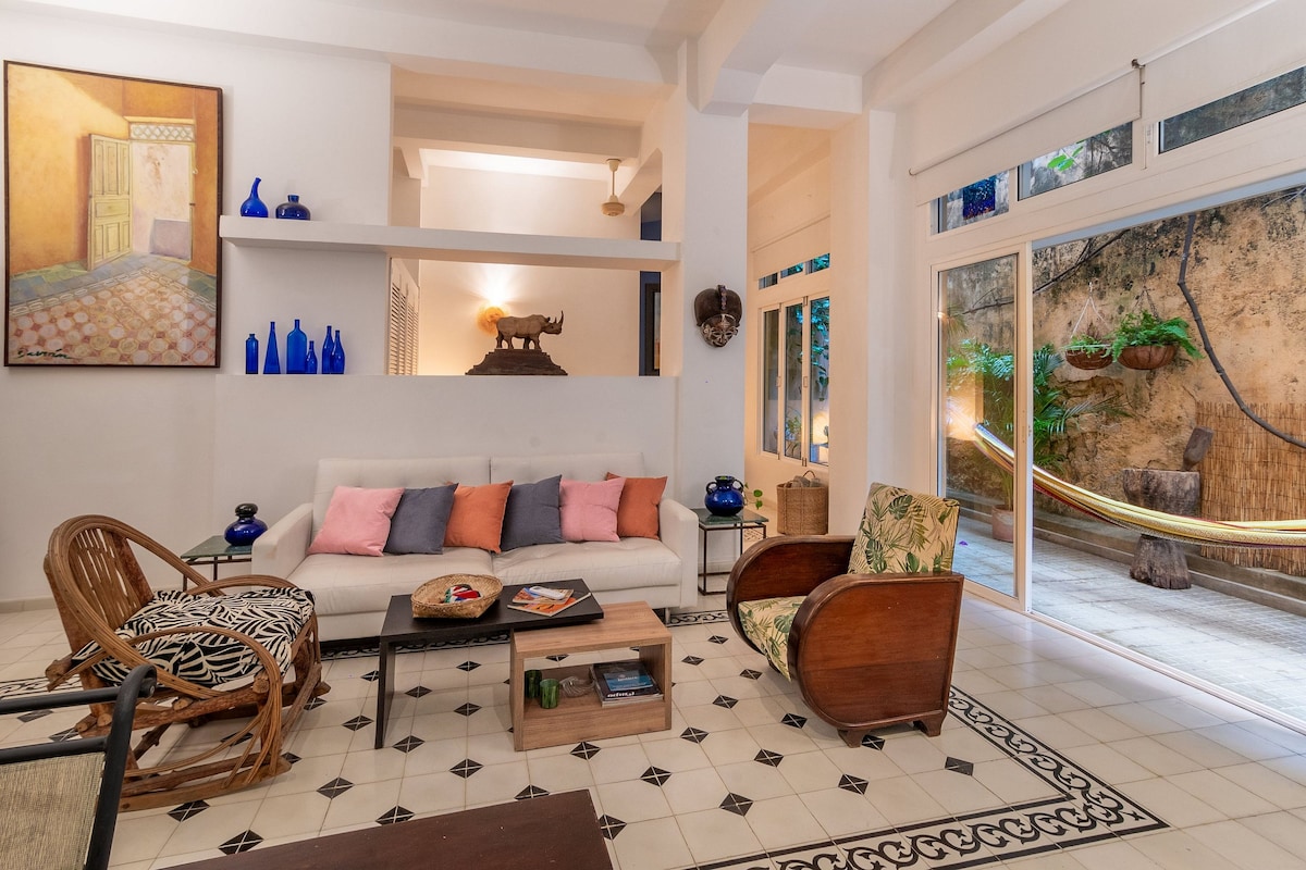 A bright living area showcases a neutral sofa adorned with various cushions. A wooden coffee table sits nearby, accompanied by vintage-style chairs. The space is complemented by large glass doors that lead to a private outdoor area featuring a hammock and greenery.