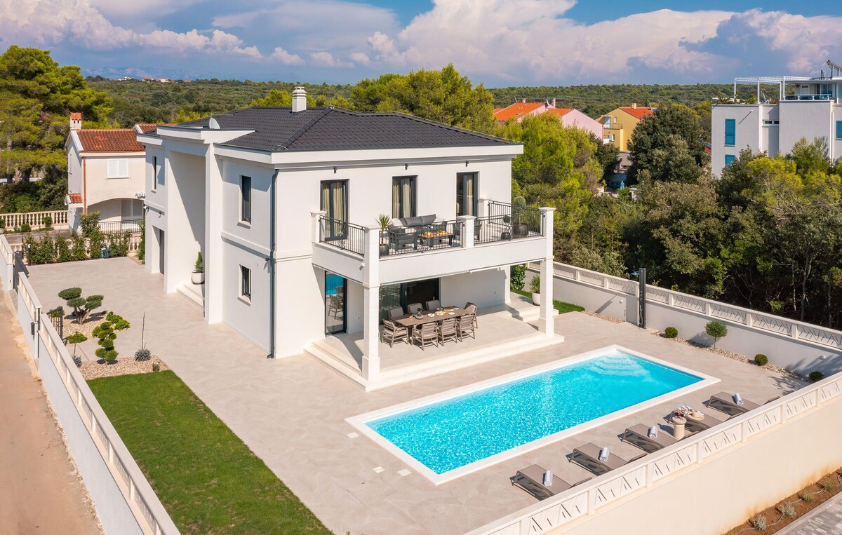 An aerial view of a modern villa features a well-maintained swimming pool surrounded by lounge chairs on a spacious terrace. The home showcases a balcony with outdoor seating, complemented by lush greenery and landscaped grounds. Clear skies provide a serene backdrop to the elegant structure.